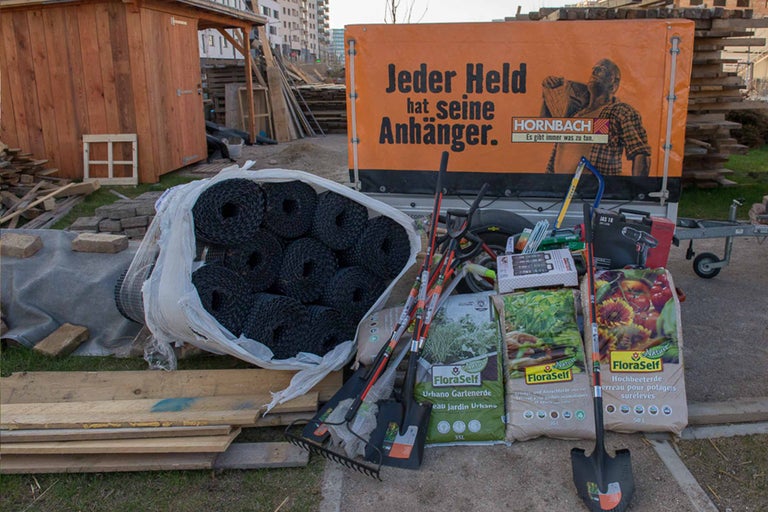 Gartenszene mit diversen Gartenprodukten wie Erde, Pflanzen und Werkzeugen vor einem Anhänger mit Hornbach Werbung