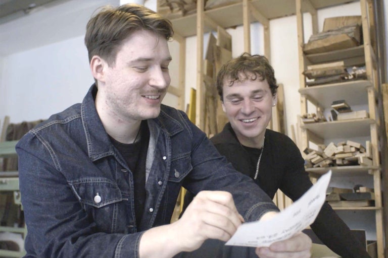 Zwei Männer betrachten ein Blatt Papier vor einem Regal mit Holz