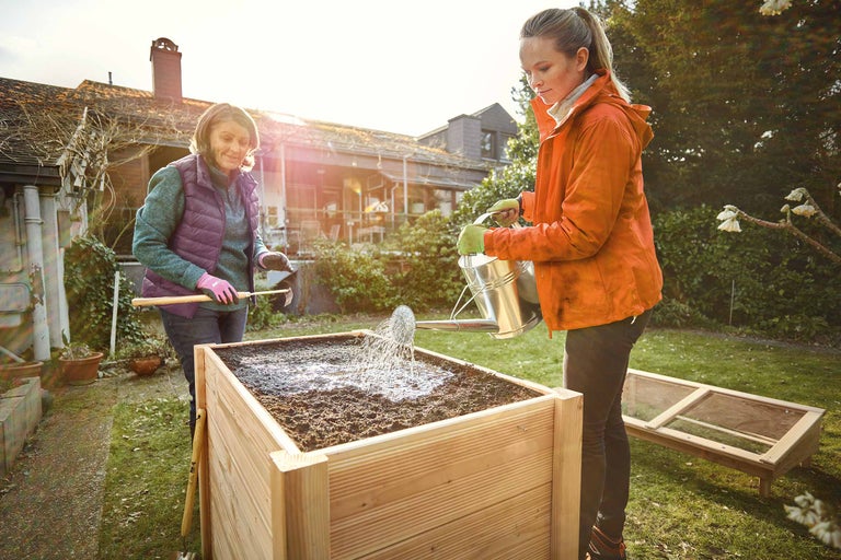 Zwei Frauen arbeiten an einem Hochbeet im Garten, eine gießt mit einer Gießkanne, die andere hält eine Harke.