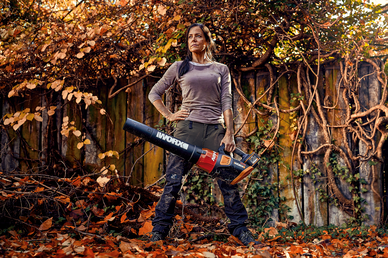 Eine Frau mit einem Worx Akku Laubbläser steht vor einem herbstlichen Gartenhintergrund.