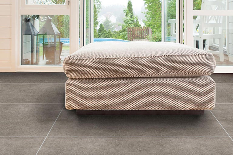 Gepolsterter Hocker auf grauem Fliesenboden vor offenem Fenster