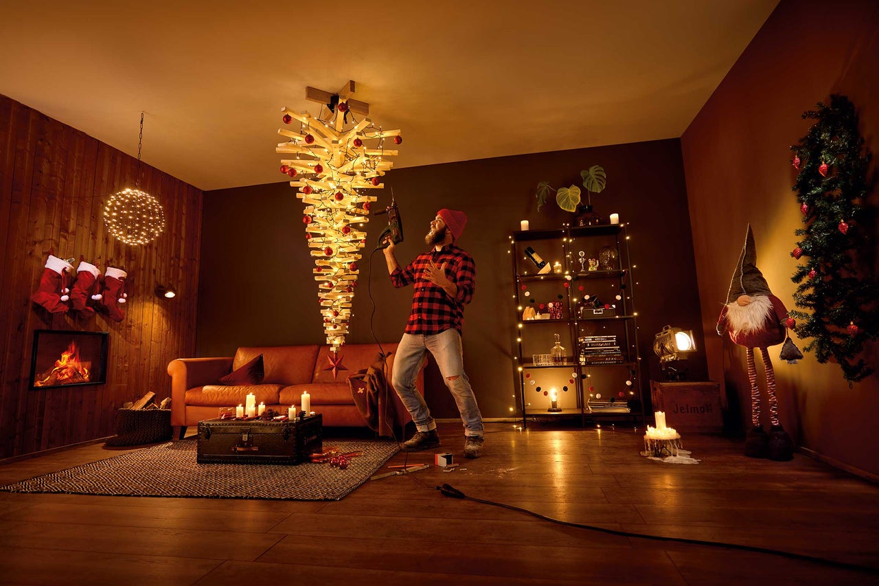 Wohnzimmer mit modernem Holzweihnachtsbaum, beleuchtet und mit roten Kugeln geschmückt, Mann mit Bohrmaschine