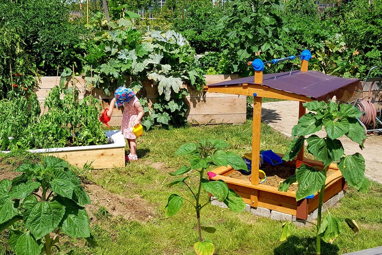 Kind gießt Pflanzen im Hochbeet neben einem Sandkasten mit Dach in einem Garten.