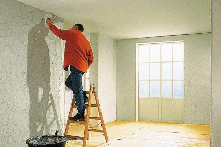 Ein Mann auf einer Leiter trägt Putz auf eine Wand auf. Werkzeuge und Eimer stehen bereit.