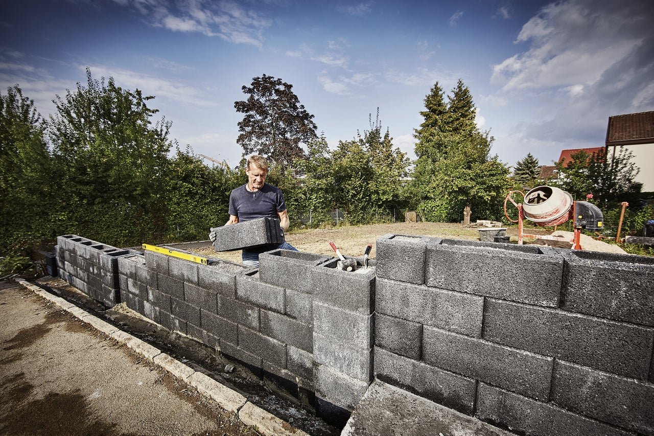 Mann baut eine Mauer aus Betonsteinen im Garten, mit Betonmischer im Hintergrund.