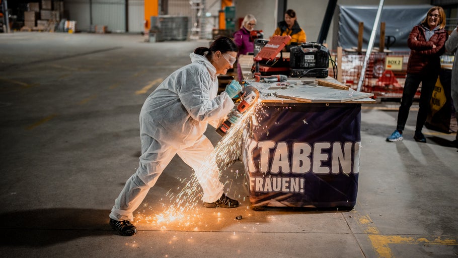 Eine Frau in Schutzanzug und Schutzbrille arbeitet mit einem Winkelschleifer, bei dem Funken fliegen.