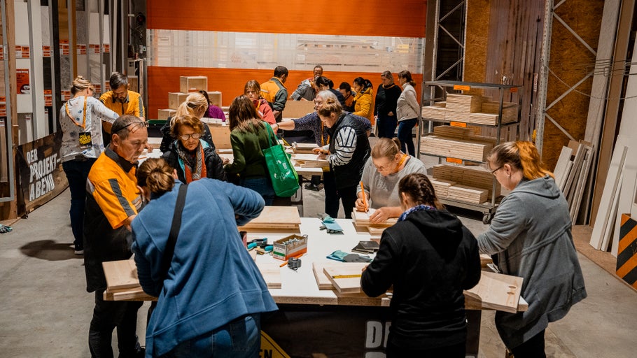 Eine Gruppe von Personen nimmt an einem Holzbearbeitungs-Workshop bei Hornbach teil und arbeitet an Tischen mit Holz und Werkzeugen.