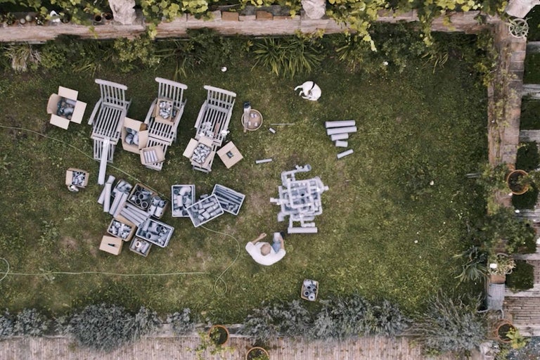 Luftaufnahme eines Gartens mit einer Person, Gartenstühlen, Pappkartons, Rohren und einem Hund
