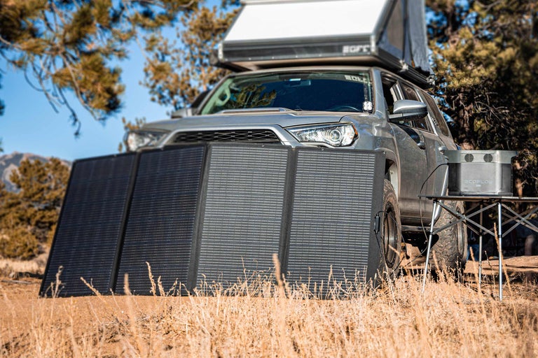 Aufnahme eines silberfarbenen Geländewagens mit Solarmodul und tragbarer Powerstation in der Natur.