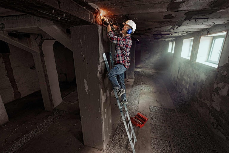 Handwerker auf einer Leiter beim Schleifen von Metall in einem Kellerraum mit einer Werkzeugkiste.