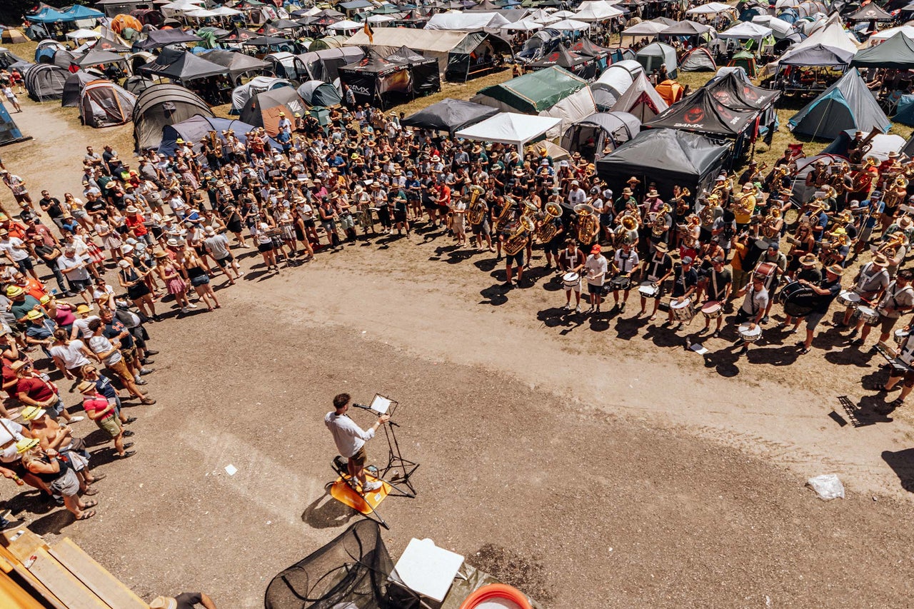 Große Menschenmenge auf einem Zeltplatz mit zahlreichen Zelten, eine Musikkapelle tritt im Freien vor Publikum auf.
