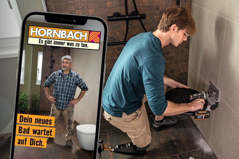 Ein Handwerker mit Beinprothese montiert einen Duschsitz an einer gefliesten Wand, während auf einem Smartphone das Hornbach Logo zu sehen ist.