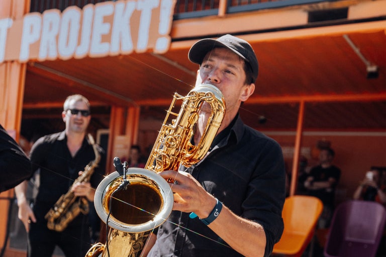 Ein Musiker spielt Baritonsaxophon vor einer orangefarbenen Konstruktion mit der Aufschrift Projekt.