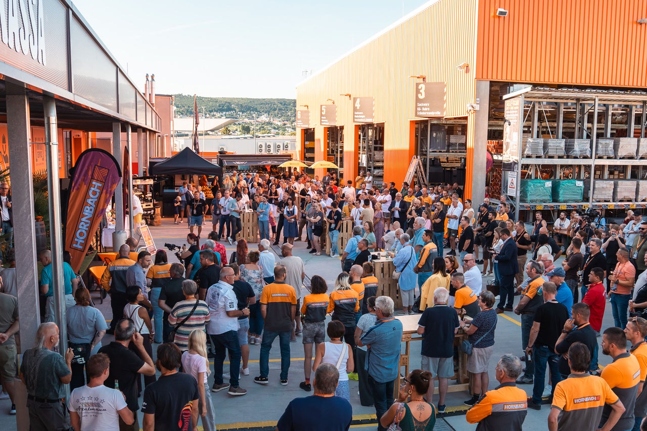 Menschenmenge bei einer hellen Außenveranstaltung vor einem Hornbach Markt. Teilnehmer tragen orange-graue Arbeitskleidung zwischen Baumaterialien.