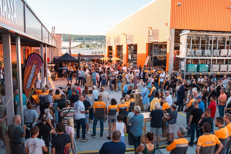 Menschenmenge bei einer hellen Außenveranstaltung vor einem Hornbach Markt. Teilnehmer tragen orange-graue Arbeitskleidung zwischen Baumaterialien.