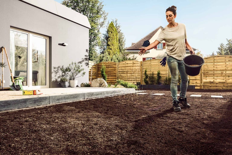 Eine Frau sät Rasensamen in einem Garten, umgeben von einem Haus und einem Holzzaun.