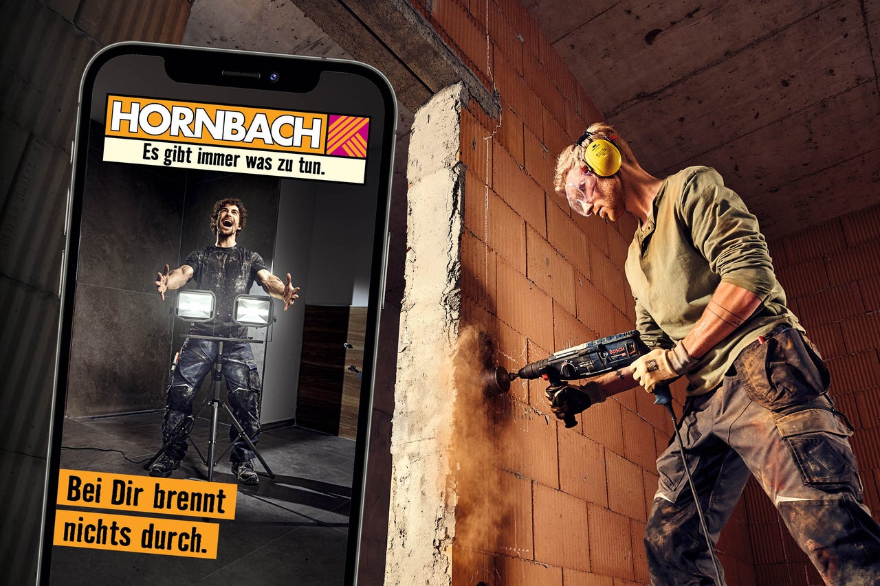 Handwerker bei der Arbeit mit einem Bohrhammer an einer Ziegelwand und Darstellung einer Hornbach App auf einem Smartphone im Vordergrund.