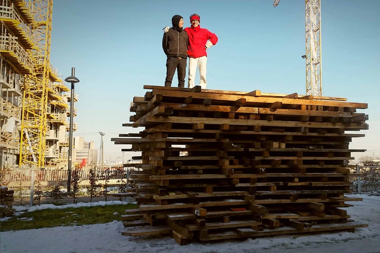 Zwei Personen stehen auf einem Stapel Holzpaletten vor einer Baustelle.