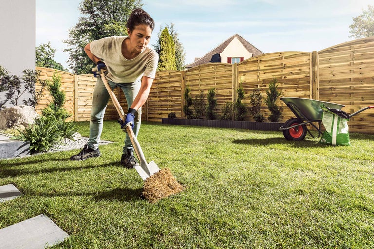 Frau arbeitet im Garten mit einem Spaten, im Hintergrund ein Zaun und eine Schubkarre