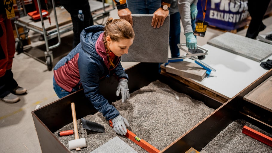 Frau verlegt Pflastersteine in einer Sandkiste mit Werkzeugen wie Wasserwaage, Hammer und Kelle.
