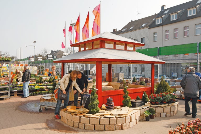 Kunden betrachten einen roten Pavillon mit Wasserspiel und Steindekoration im Hornbach Gartencenter.