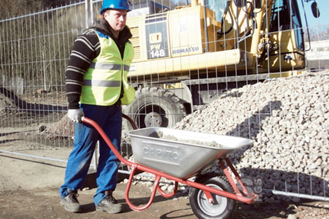 Ein Bauarbeiter schiebt eine Schubkarre mit Schutt vor einem Bagger auf einer Baustelle.