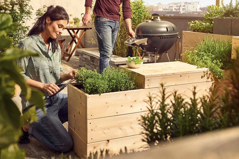 Szene auf einer Dachterrasse mit Hochbeet aus Holz, Grill und zwei Personen, die sich mit Gartenarbeit und Grillen beschäftigen.