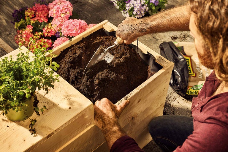 Eine Person befüllt einen hölzernen Pflanzkasten mit Erde unter Verwendung einer kleinen Schaufel.