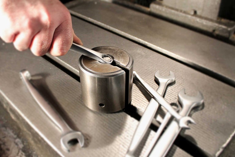 Eine Hand bedient einen Maulschlüssel an einem Metallzylinder, umgeben von weiteren Schraubenschlüsseln auf einer Werkbank.