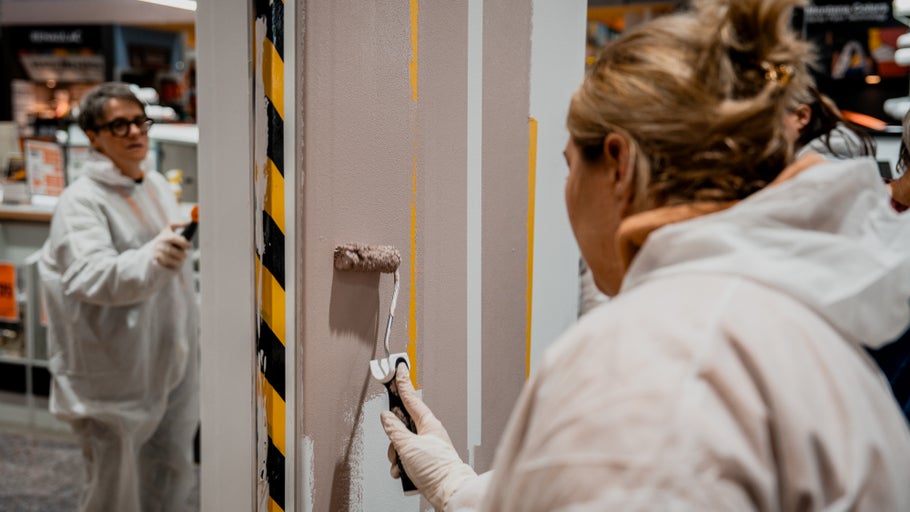 Personen streichen eine Wand mit einem Farbroller, wobei gelbe Streifen sichtbar sind. Maleranzüge werden getragen.