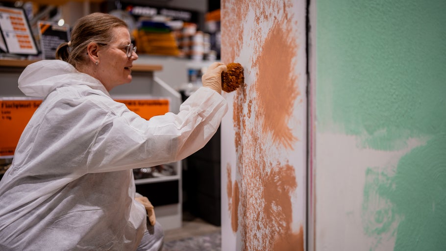 Frau im Schutzanzug trägt Farbe mit einem Schwamm auf eine Wand auf