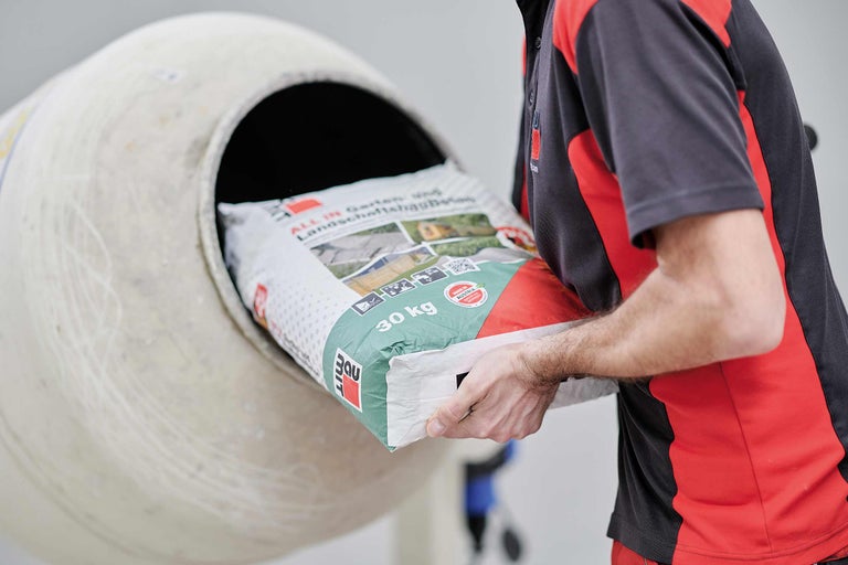 Ein Mann leert einen 30 Kilogramm Sack All-in-Garten- und Landschaftsbau-Beton in einen Betonmischer.