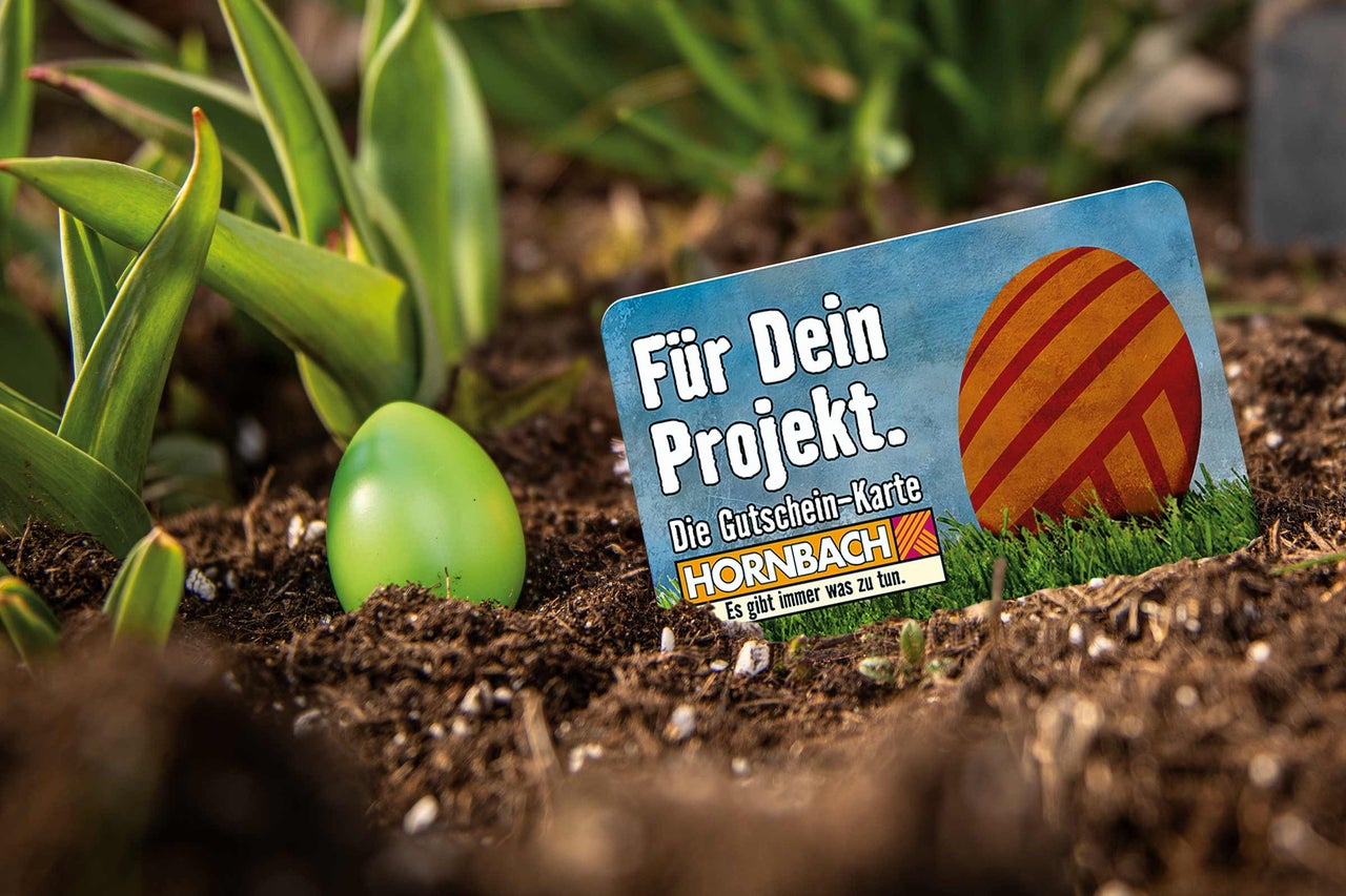 Hornbach Gutscheinkarte mit Osterei Motiv und Hornbach Logo in einem Gartenbeet mit Erde, Pflanzen und einem dekorativen Ei.