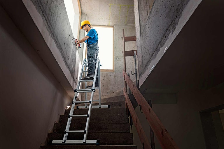 Mann mit Bohrmaschine auf einer Mehrzweckleiter in einem Treppenhaus