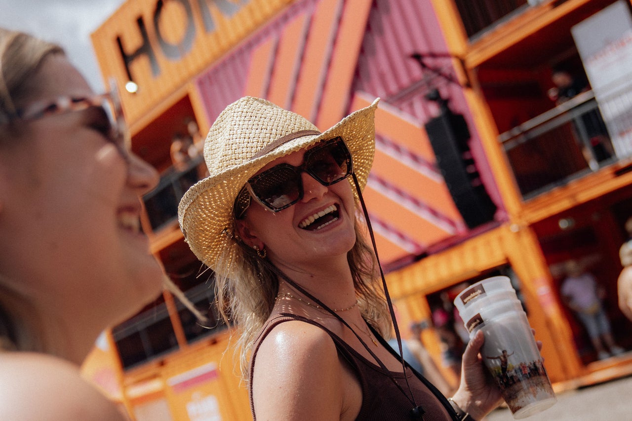 Frau mit Strohhut und Sonnenbrille lächelt auf einem Festival vor einem Gebäude aus Containern mit Hornbach Logo.