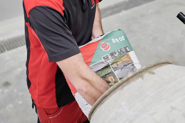 Eine Person leert einen 30 Kilogramm Sack Estrichmörtel in einen Betonmischer.