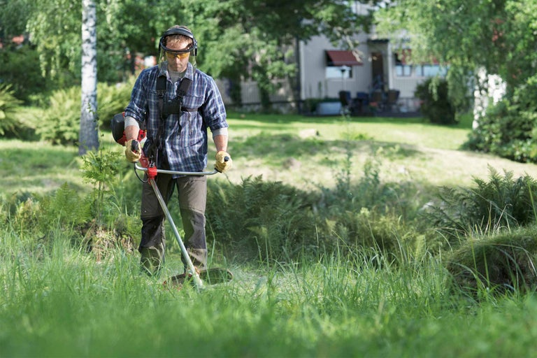 Ein Mann mit Schutzkleidung mäht mit einer Motorsense das Gras in einem Garten.