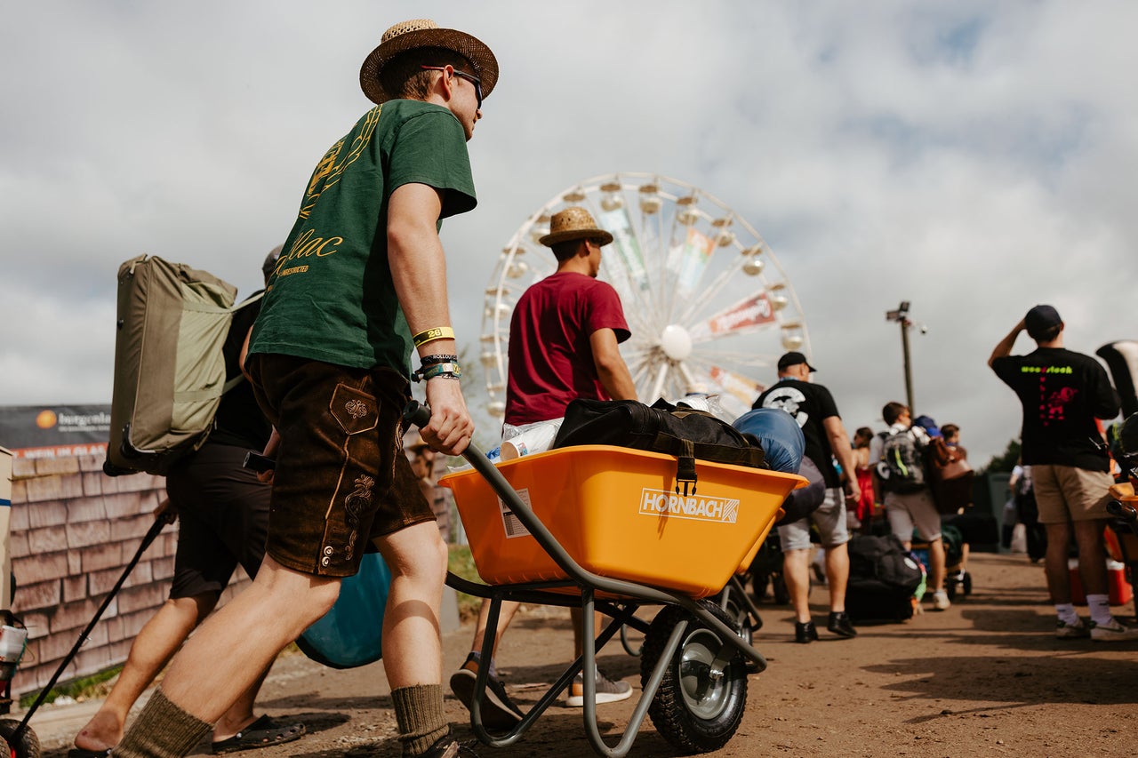 Mann schiebt eine mit Campingausrüstung beladene Schubkarre auf einem Festivalgelände, im Hintergrund ein Riesenrad. Hornbach Logo.