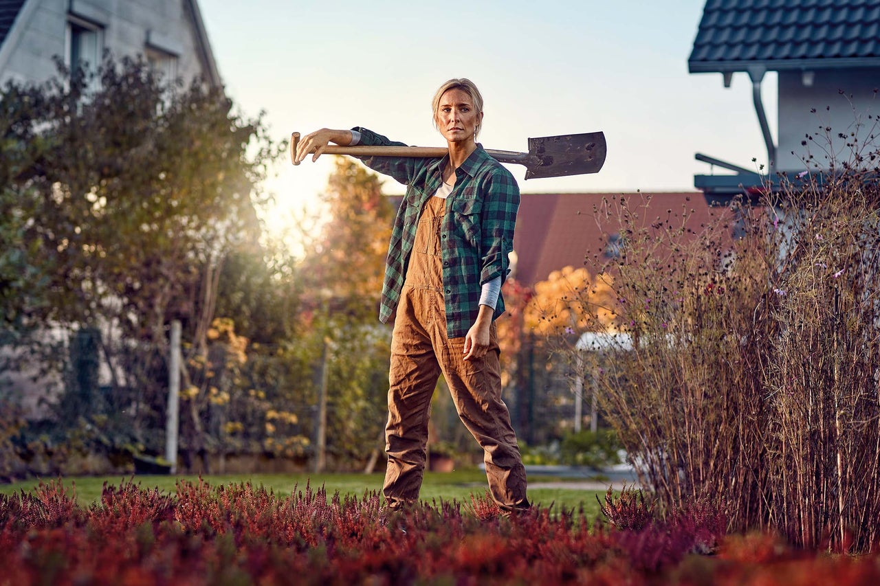 Frau in Gartenkleidung steht mit einem Spaten auf der Schulter in einem Garten vor einem Haus bei Sonnenuntergang.