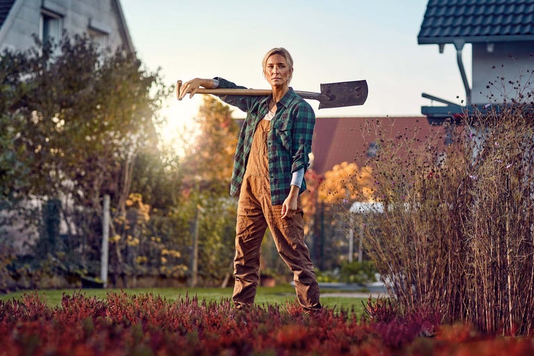 Frau in Gartenkleidung steht mit einem Spaten auf der Schulter in einem Garten vor einem Haus bei Sonnenuntergang.
