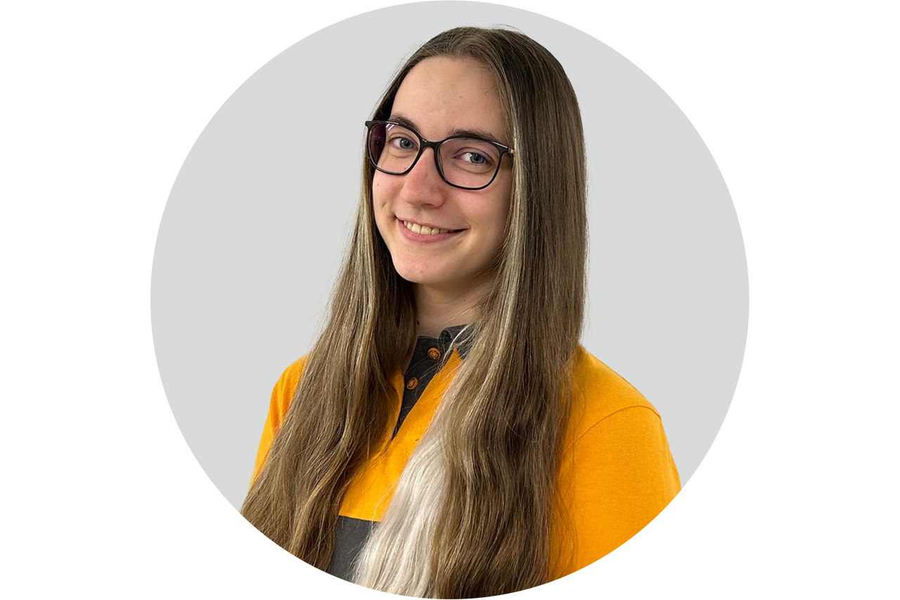 Porträt einer lächelnden jungen Frau mit Brille und langem Haar, die ein gelbes Oberteil trägt.