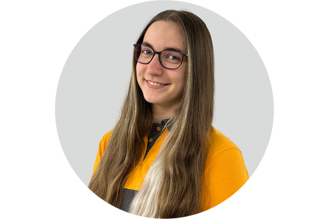 Porträt einer lächelnden jungen Frau mit Brille und langem Haar, die ein gelbes Oberteil trägt.