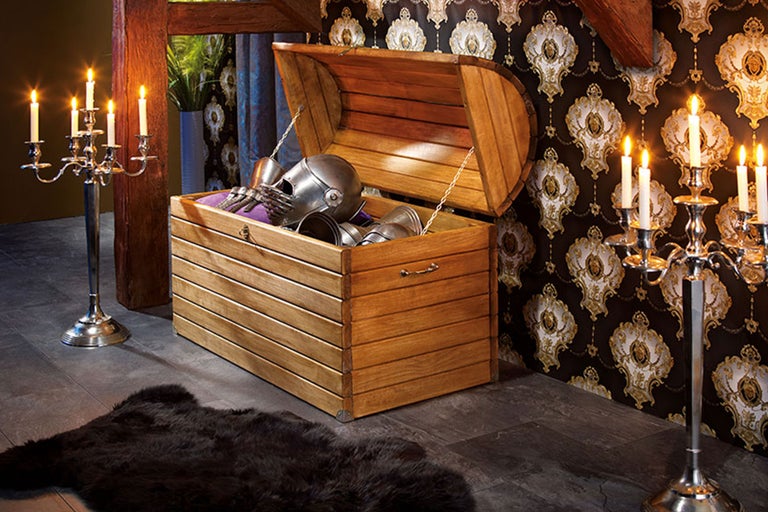 Szene mit offener Holztruhe gefüllt mit Ritterrüstung, umgeben von silbernen Kerzenständern und einem dunklen Teppich