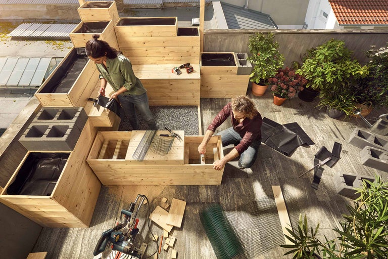 Zwei Personen bauen auf einer Dachterrasse ein Hochbeet aus Holz, Pflanzfolie, Steinen und Werkzeug.