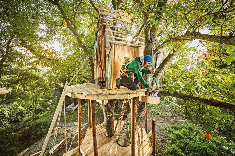 Eine Person arbeitet an einem Baumhaus in einem Baum mit grünen Blättern.