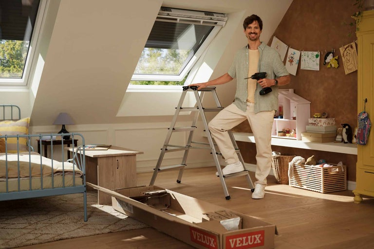 Mann mit Bohrmaschine auf einer Leiter vor einem Dachfenster in einem Kinderzimmer