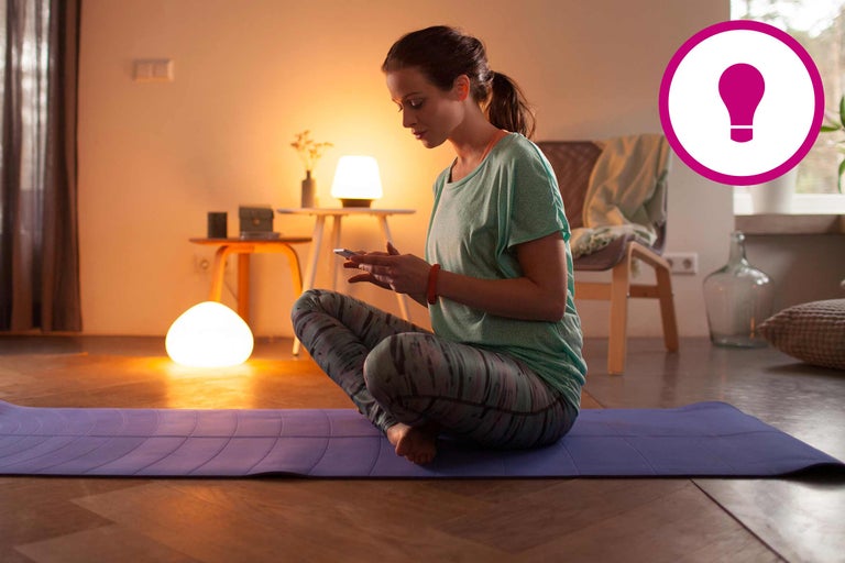 Frau sitzt auf einer Yogamatte und benutzt ein Smartphone in einem Wohnzimmer mit Tischlampen.