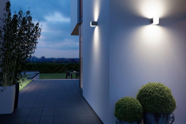 Beleuchtete Außenansicht mit zwei quadratischen Wandleuchten, Bepflanzung und Terrassenbereich.