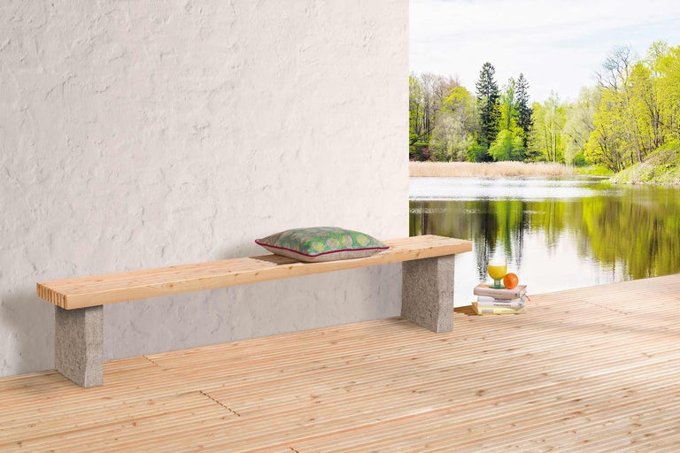 Holzbank mit Steinfüßen, Kissen, Getränk und Büchern auf einer Holzterrasse mit Seeblick