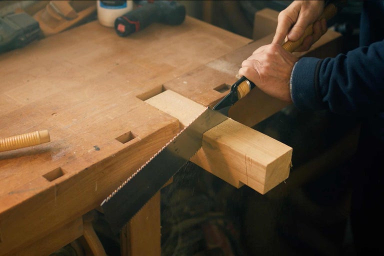 Eine Person sägt ein Stück Holz auf einer Hobelbank mit einer Handsäge.